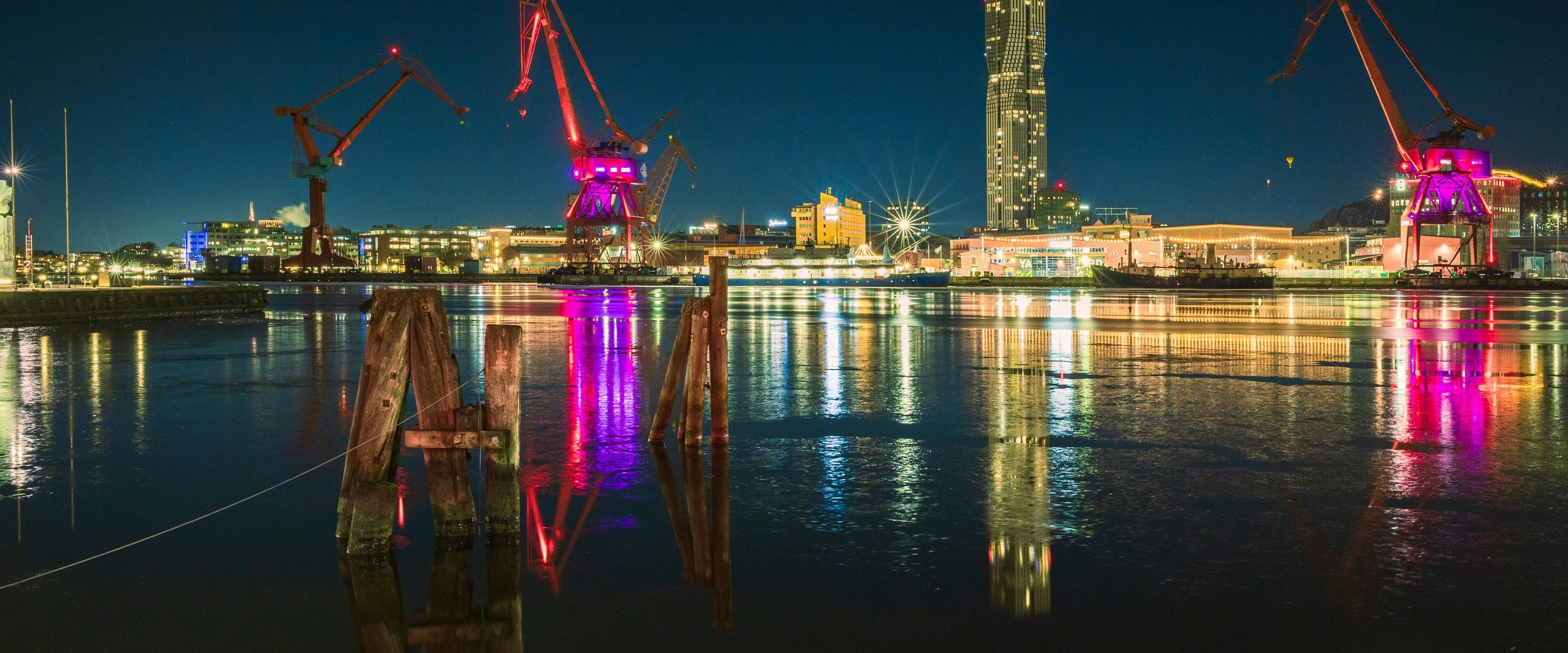 A harbor at night, with red-lit cranes and high-rise buildings reflected on the water